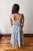 A woman viewed from behind wearing a sleeveless, tiered midi dress with a blue floral pattern on a white background, standing on a wooden floor against a plain white wall.