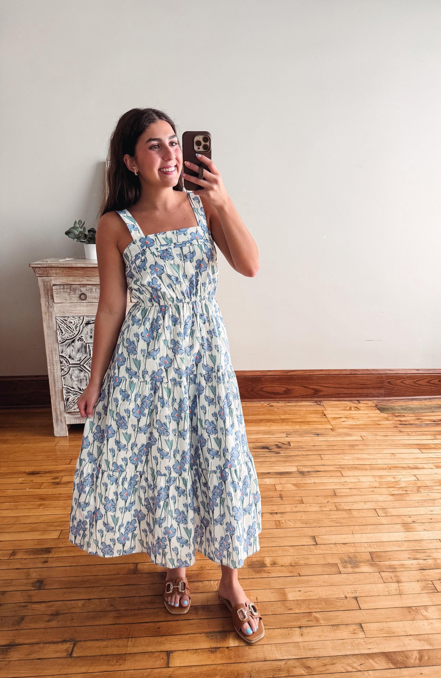 A woman takes a mirror selfie wearing a sleeveless midi dress with a blue and white floral pattern, standing on a wooden floor in front of a plain wall.