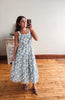 A woman takes a mirror selfie wearing a sleeveless midi dress with a blue and white floral pattern, standing on a wooden floor in front of a plain wall.
