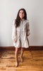 A woman stands in a room with a light gray wall and wooden floor, wearing a cream-colored long-sleeve dress with a subtle floral pattern and light blue trim, paired with tan knee-high boots.