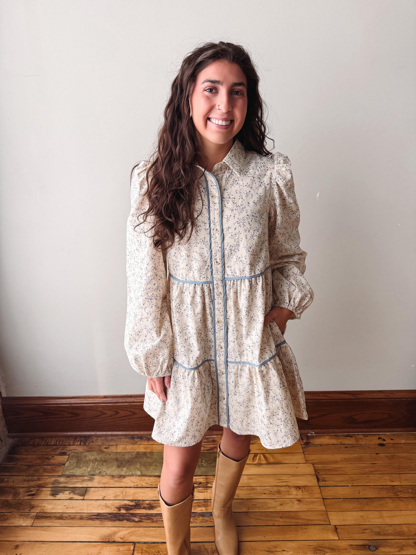 A woman wearing a cream-colored floral shirt dress with light blue trim and long sleeves, paired with tan knee-high boots, standing in a room with a white wall and wooden floor.