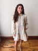A woman wearing a cream-colored floral shirt dress with light blue trim and long sleeves, paired with tan knee-high boots, standing in a room with a white wall and wooden floor.