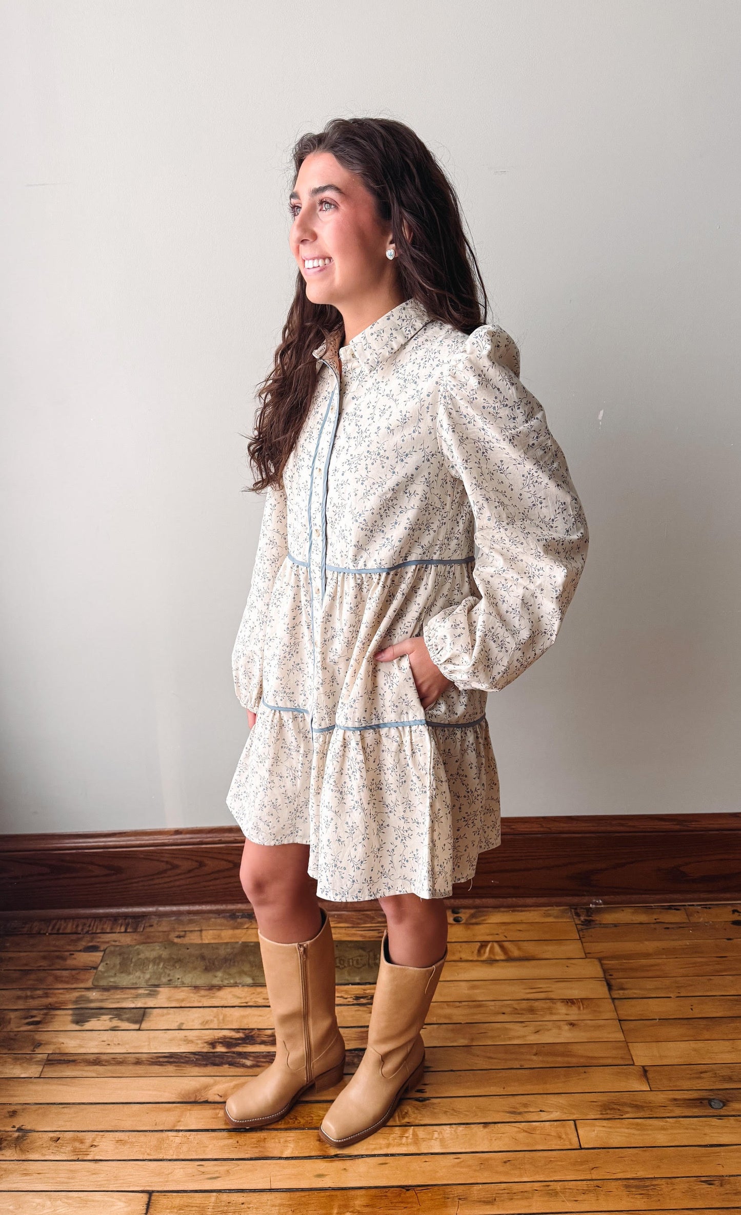 A woman modeling a cream-colored, long-sleeved mini dress with a delicate floral pattern and light blue piping, paired with tan knee-high boots, standing on a wooden floor against a plain wall.