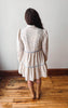 A woman stands with her back to the camera wearing a cream-colored long-sleeve dress with a delicate blue floral pattern and light blue trim, paired with tan knee-high boots, set against a plain wall and wooden floor.