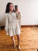 A woman takes a mirror selfie wearing a cream-colored floral print shirt dress with light blue piping and beige knee-high boots, standing on a wooden floor against a white wall.
