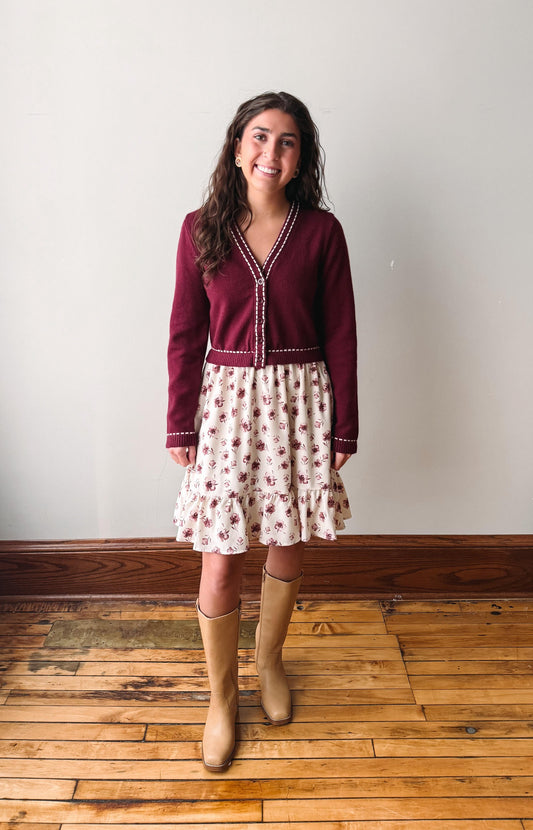 A woman stands in a room with a white wall and wooden floor, wearing a burgundy cardigan with white trim over a cream tiered skirt with a red floral print, paired with tan knee-high boots.