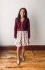 A woman stands in a room with a white wall and wooden floor, wearing a burgundy cardigan with white trim over a cream tiered skirt with a red floral print, paired with tan knee-high boots.