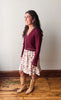 A woman stands in profile wearing a burgundy knit cardigan with white trim over a cream floral print skirt, paired with tan knee-high boots, against a plain light gray wall and wooden floor.