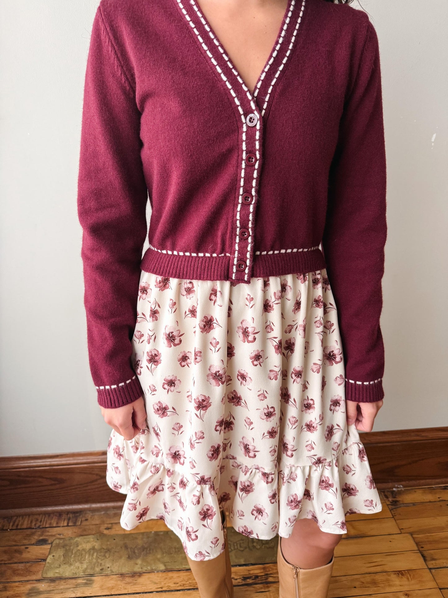 A person wearing a burgundy cardigan with white contrast stitching over a cream tiered skirt with a pink floral print, paired with tan boots, standing on a wooden floor against a plain wall.