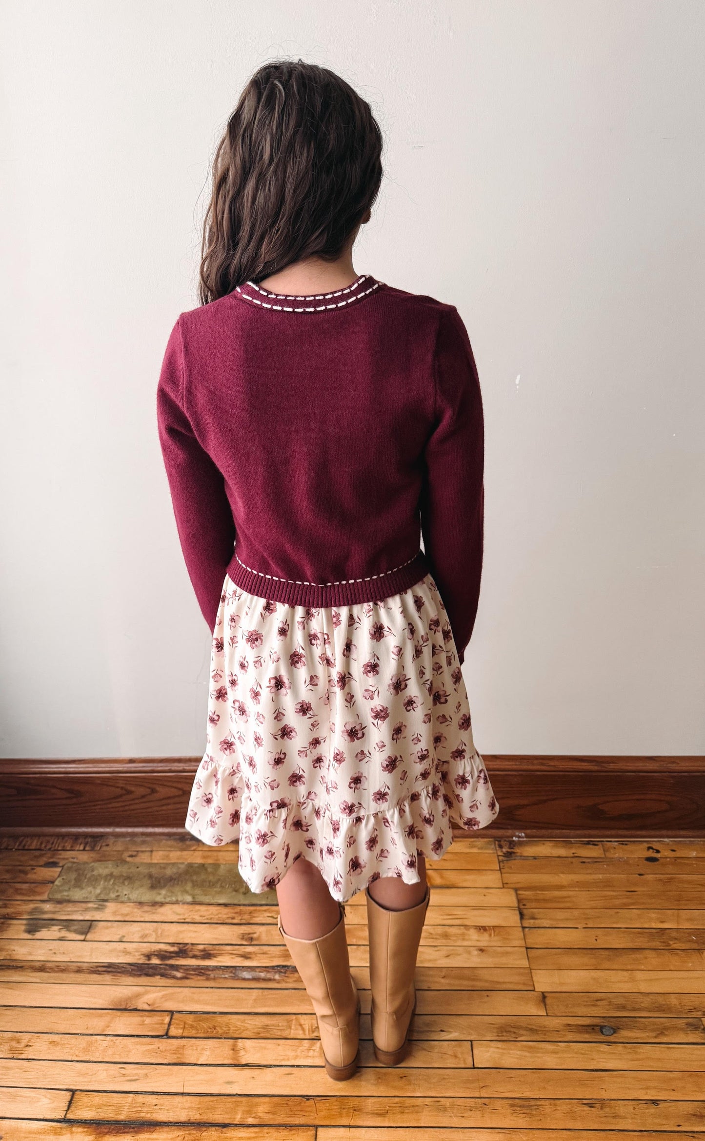 A woman viewed from behind wearing a burgundy knit cardigan and a cream floral skirt, standing on a wooden floor against a plain wall.
