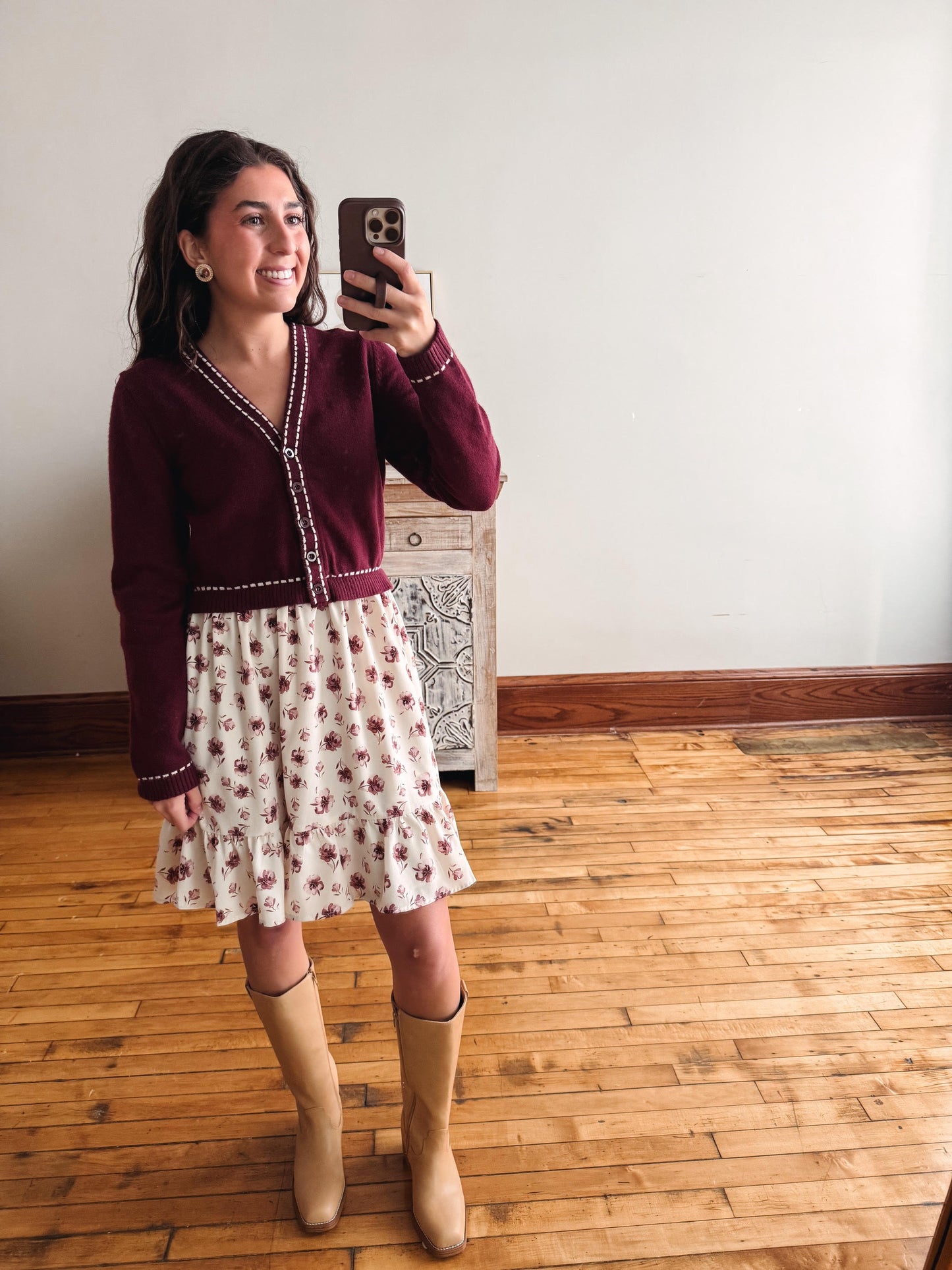 A woman takes a mirror selfie wearing a burgundy knit cardigan with white trim, a cream floral skirt, and tan knee-high boots in a room with wooden floors and a white wall.