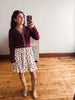 A woman takes a mirror selfie wearing a burgundy knit cardigan with white trim, a cream floral skirt, and tan knee-high boots in a room with wooden floors and a white wall.