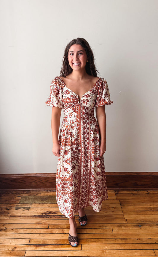 A woman wearing a bohemian-style midi dress with a rust red and cream pattern, standing on a wooden floor against a plain off-white wall.