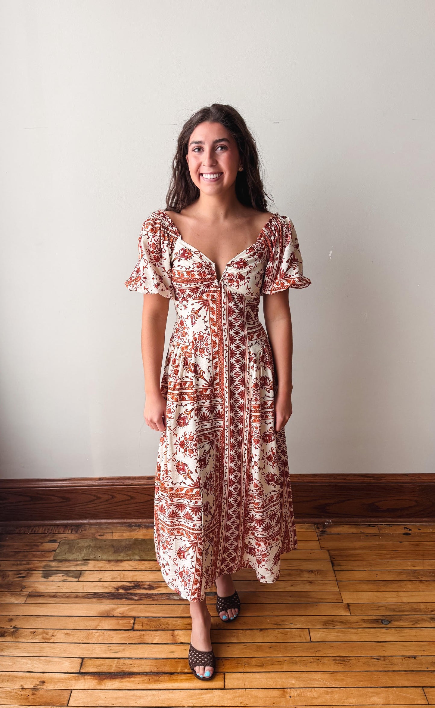 A woman wearing a bohemian-style midi dress with a rust red and cream pattern, standing on a wooden floor against a plain off-white wall.