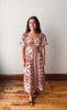 A woman wearing a bohemian-style midi dress with a rust red and cream pattern, standing on a wooden floor against a plain off-white wall.