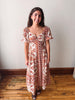 A woman modeling a bohemian-style midi dress with a rust orange and cream pattern, standing on a wooden floor against a plain off-white wall.