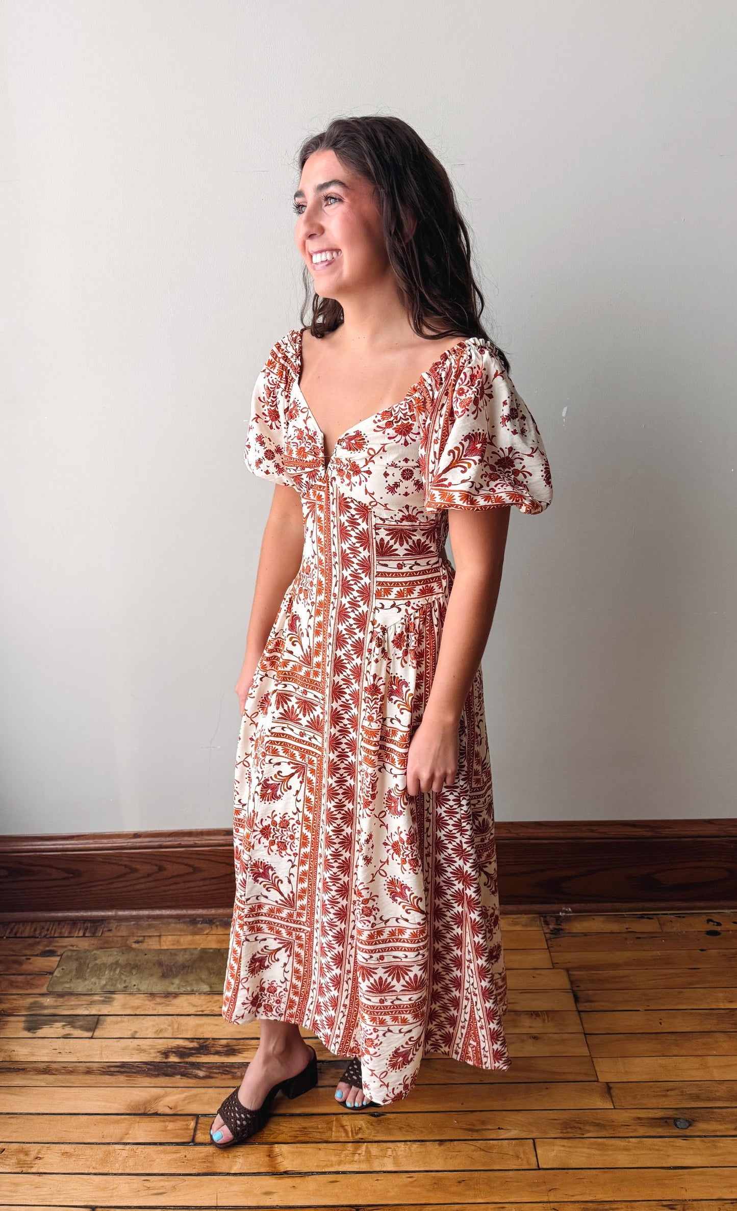 A woman modeling a bohemian-style maxi dress with puff sleeves, a V-neckline, and a rust-orange and cream pattern, standing on a wooden floor against a plain light gray wall.