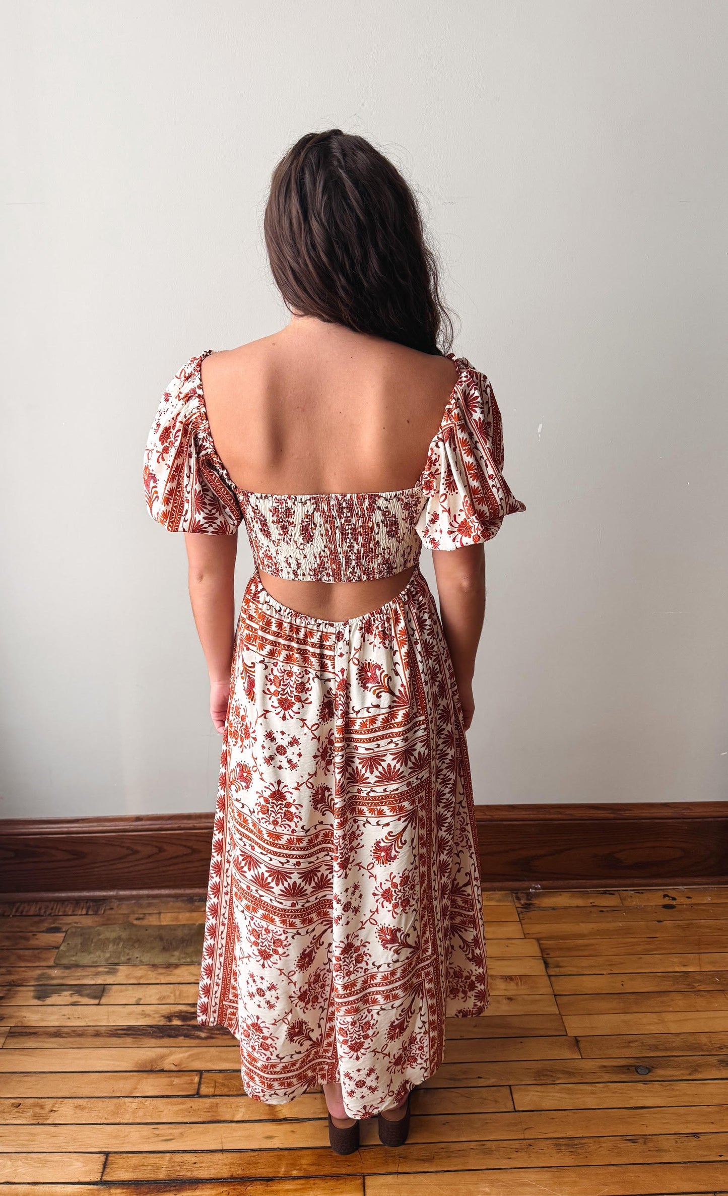 A woman viewed from behind wearing a two-piece bohemian-style dress set with a rust red and cream pattern, standing on a wooden floor against a plain off-white wall.