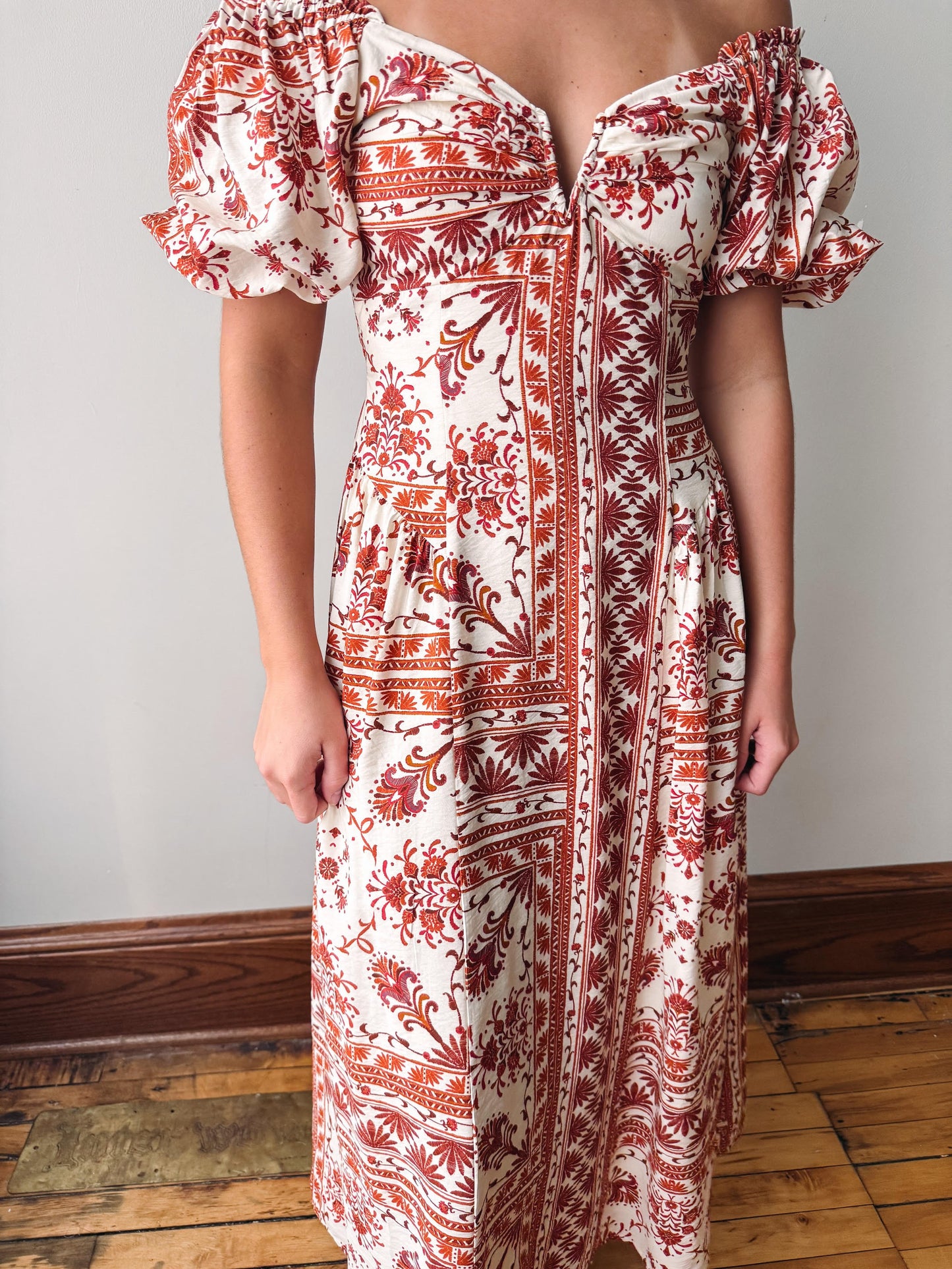 A woman wearing a cream-colored off-the-shoulder midi dress with a rust red botanical and geometric pattern, standing in front of a plain wall and wooden floor.