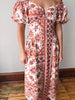 A woman wearing a cream-colored off-the-shoulder midi dress with a rust red botanical and geometric pattern, standing in front of a plain wall and wooden floor.