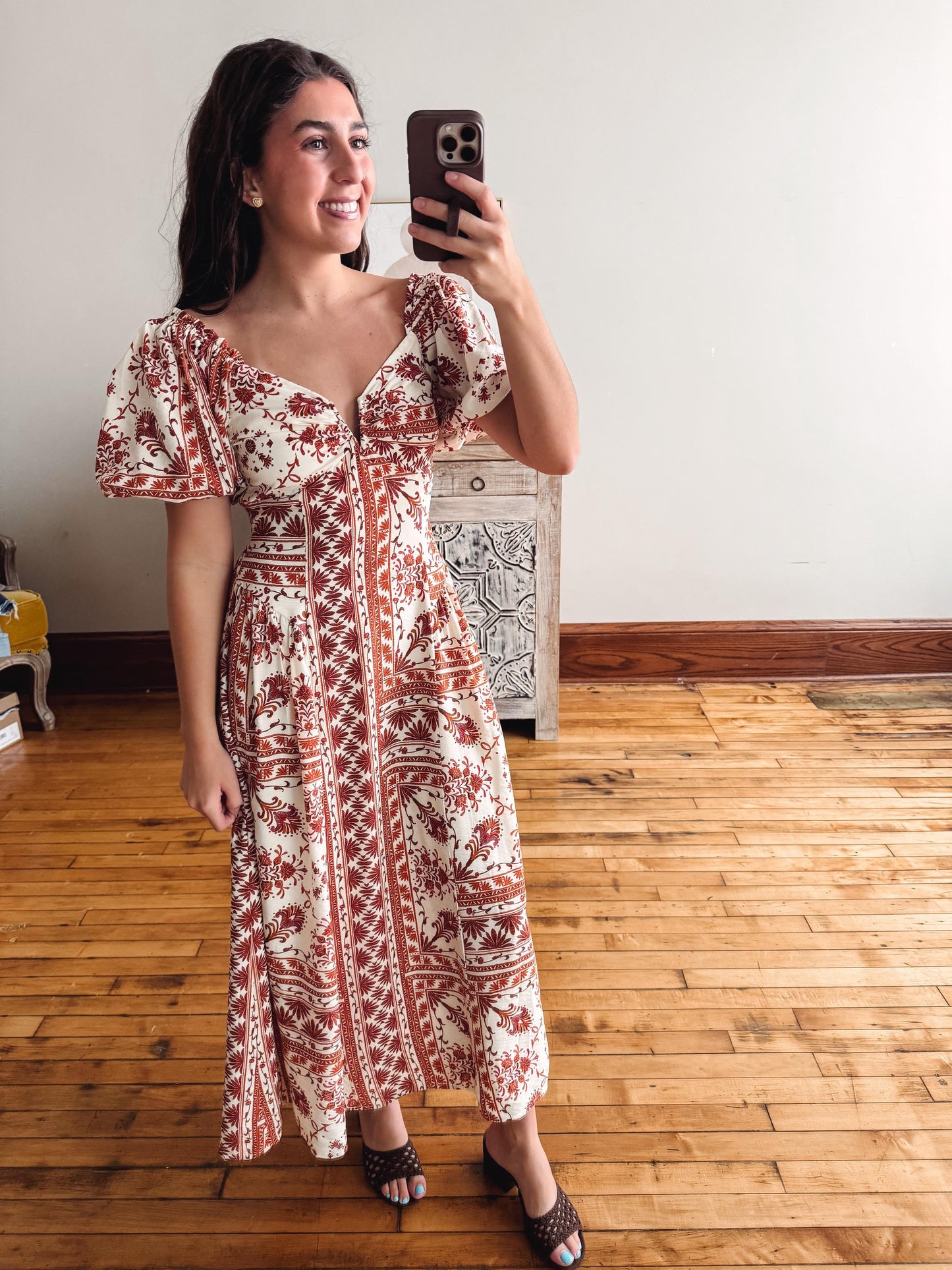 A woman takes a mirror selfie wearing a short-sleeve midi dress with a rust red and cream geometric pattern. She is standing in a room with hardwood floors and a white wall.