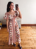 A woman takes a mirror selfie wearing a short-sleeve midi dress with a rust red and cream geometric pattern. She is standing in a room with hardwood floors and a white wall.
