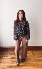 A woman modeling a black long-sleeve blouse with a white floral pattern and brown wide-leg pants, standing on a wooden floor against a plain off-white wall.