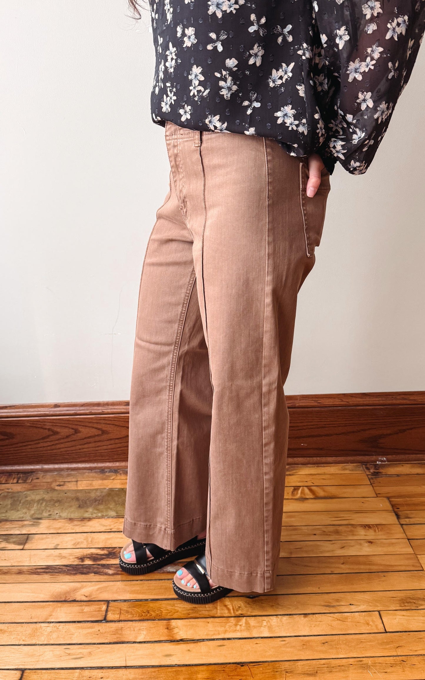 A person wearing wide-leg taupe pants and a black floral long-sleeve top, standing on a wooden floor against a white wall.