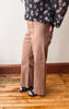 A person wearing wide-leg taupe pants and a black floral long-sleeve top, standing on a wooden floor against a white wall.