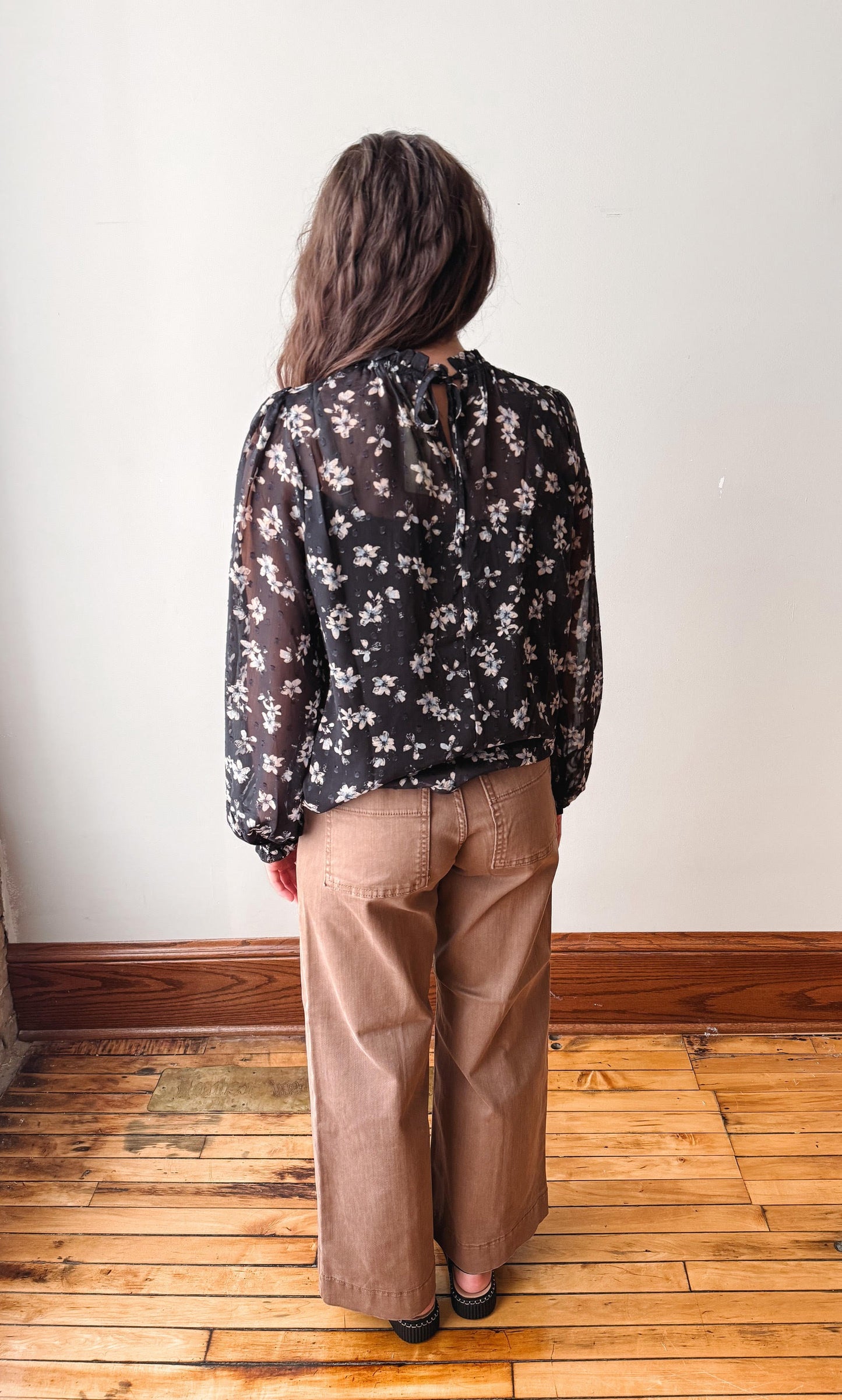 A woman viewed from behind wearing a sheer black long-sleeve blouse with a white floral pattern and brown wide-leg pants, standing on a wooden floor against a plain white wall.