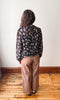 A woman viewed from behind wearing a sheer black long-sleeve blouse with a white floral pattern and brown wide-leg pants, standing on a wooden floor against a plain white wall.