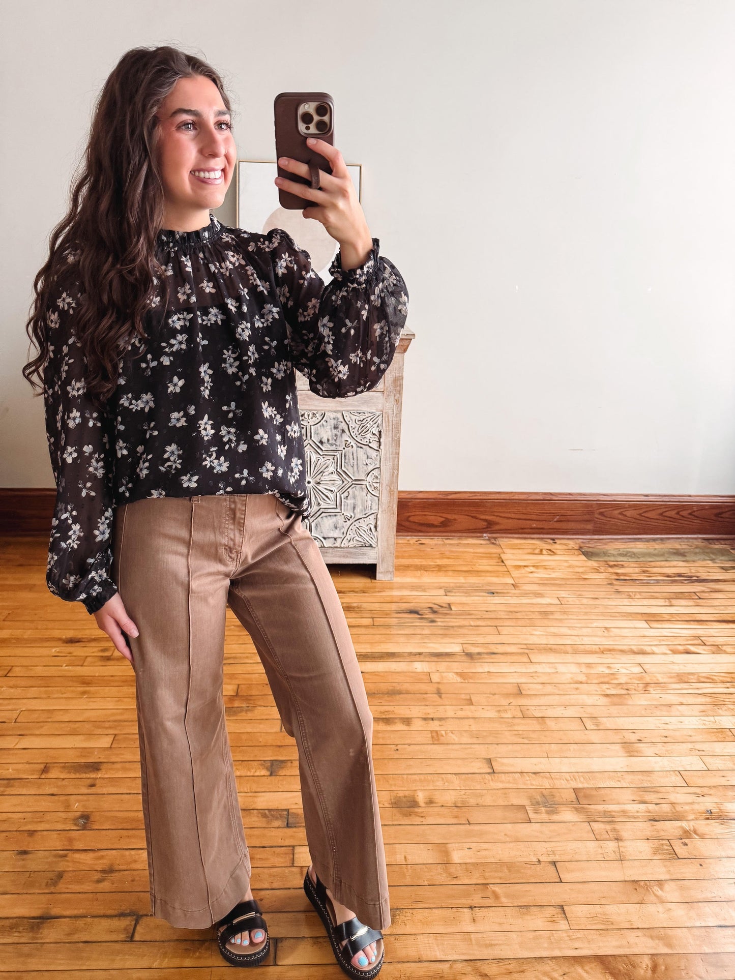 A woman takes a mirror selfie in a room with hardwood floors and a white wall, wearing a black long-sleeve blouse with a white floral pattern and brown wide-leg pants.