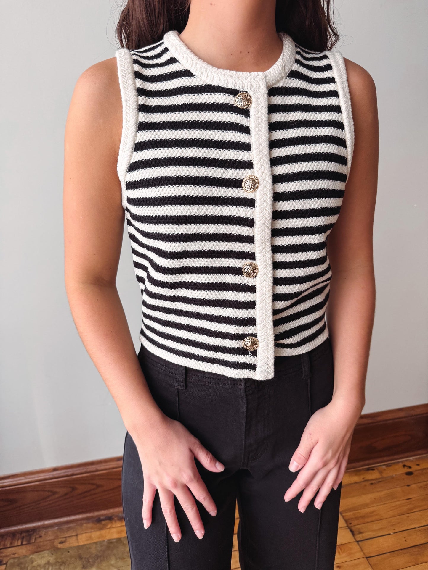 A sleeveless knit vest with black and white horizontal stripes, featuring a cream-colored trim and a front button closure, worn by a model with black pants.