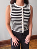 A sleeveless knit vest with black and white horizontal stripes, featuring a cream-colored trim and a front button closure, worn by a model with black pants.