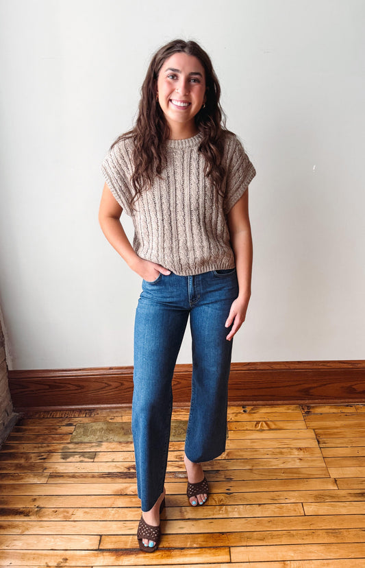 A woman stands indoors wearing a beige short-sleeve cable-knit sweater, blue jeans, and brown patterned heels against a plain off-white wall with wood trim and a wooden floor.