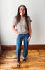 A woman stands indoors wearing a beige short-sleeve cable-knit sweater, blue jeans, and brown patterned heels against a plain off-white wall with wood trim and a wooden floor.