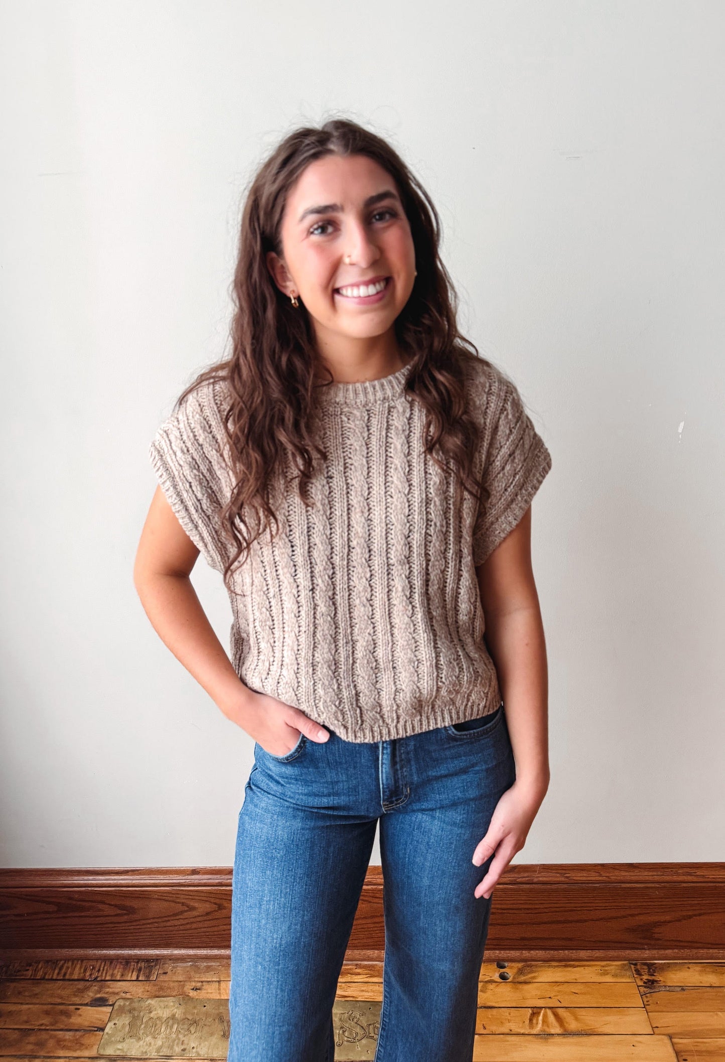 A woman with long brown hair stands indoors wearing a beige short-sleeve ribbed knit top and blue jeans against a plain off-white wall with a wooden baseboard and floor.