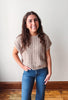 A woman with long brown hair stands indoors wearing a beige short-sleeve ribbed knit top and blue jeans against a plain off-white wall with a wooden baseboard and floor.