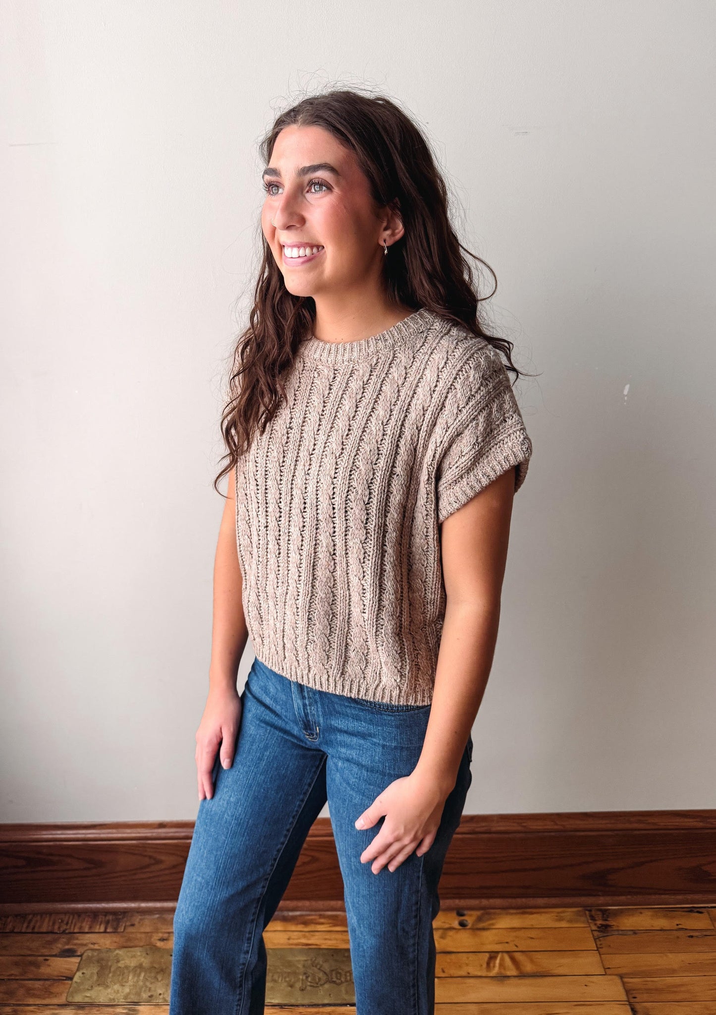 A woman with long brown hair stands in a room wearing a beige cable-knit short-sleeve top and blue jeans, posing against a plain off-white wall with a wooden baseboard and floor.