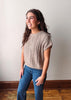 A woman with long brown hair stands in a room wearing a beige cable-knit short-sleeve top and blue jeans, posing against a plain off-white wall with a wooden baseboard and floor.