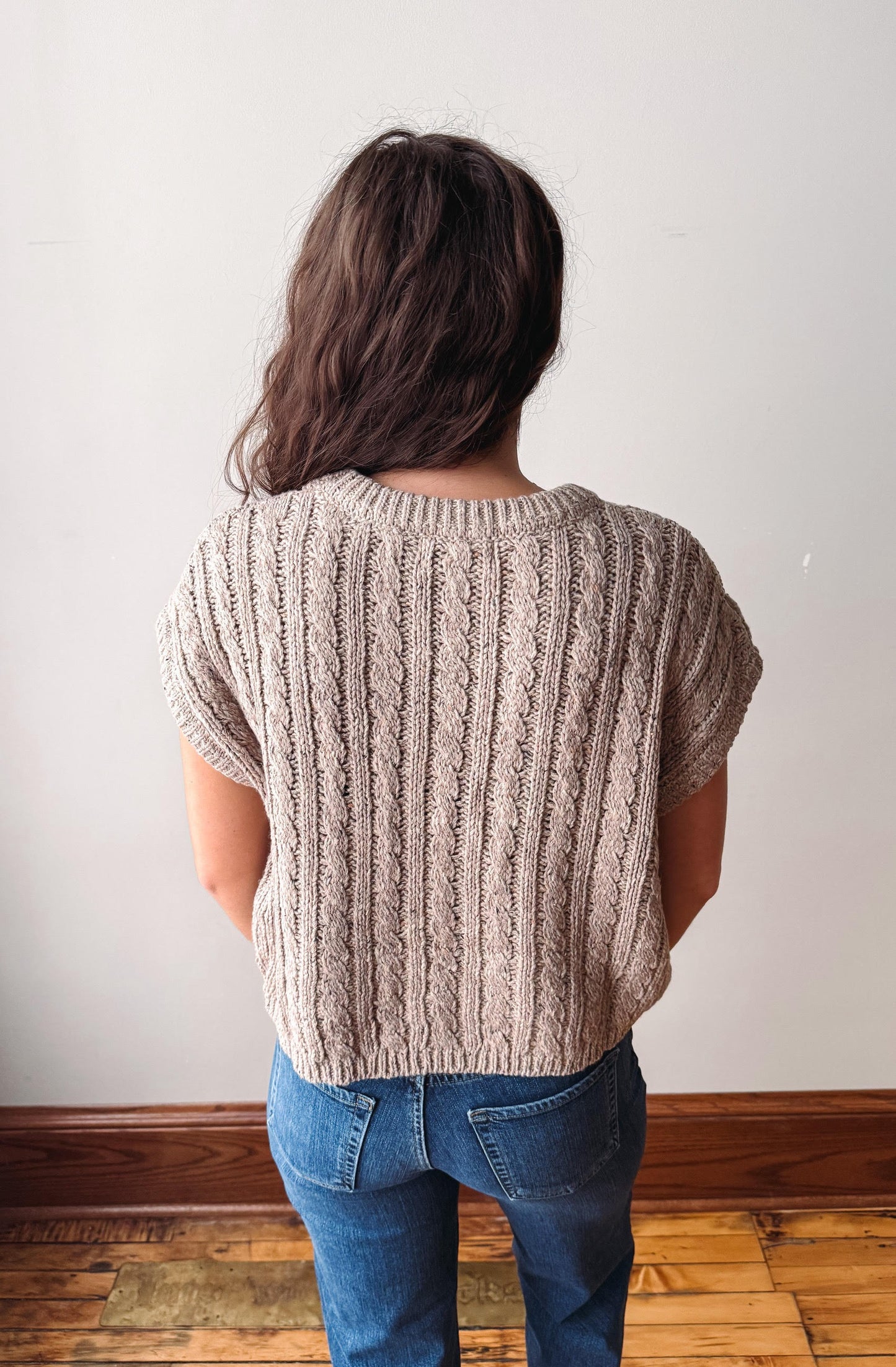 A person viewed from behind wearing a beige short-sleeve cable-knit sweater and blue jeans, standing against a plain off-white wall with a wooden baseboard and floor.