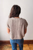 A person viewed from behind wearing a beige short-sleeve cable-knit sweater and blue jeans, standing against a plain off-white wall with a wooden baseboard and floor.