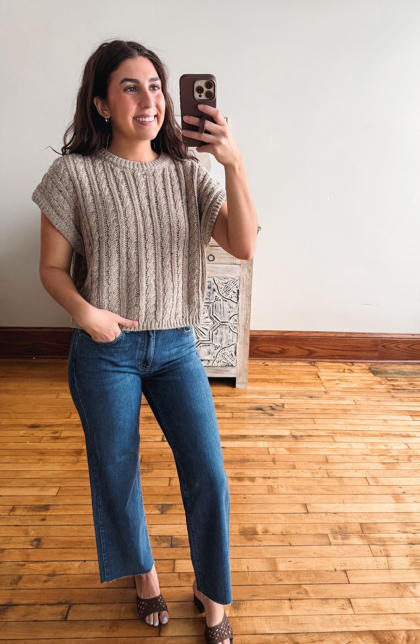 A woman takes a mirror selfie wearing a beige short-sleeve cable-knit sweater and blue jeans in a room with a light-colored wall and wooden floor.