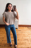 A woman takes a mirror selfie wearing a beige short-sleeve cable-knit sweater and blue jeans in a room with a light-colored wall and wooden floor.