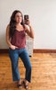 A woman takes a mirror selfie wearing a sleeveless dusty rose camisole top and blue cropped jeans in a room with light wood floors and a white wall.