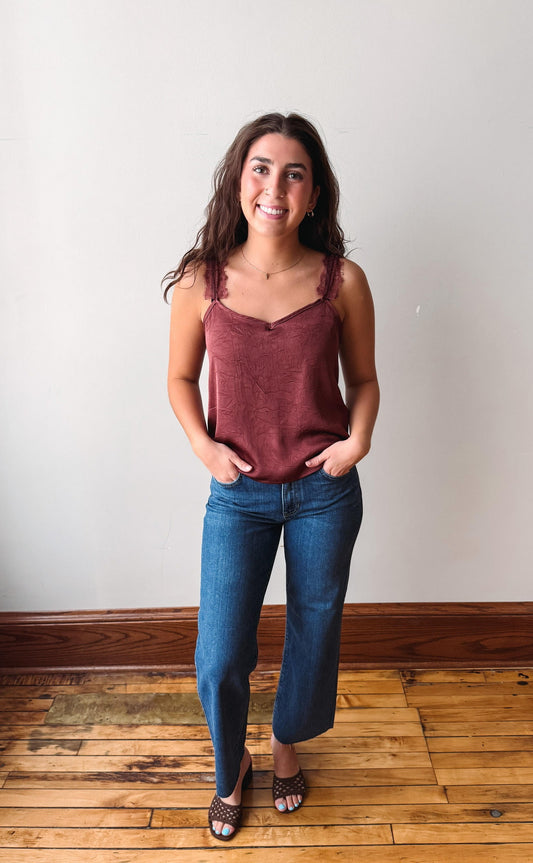 A woman stands indoors wearing a sleeveless dusty rose camisole top and blue jeans, with her hands in her pockets, against a plain off-white wall and wooden floor.