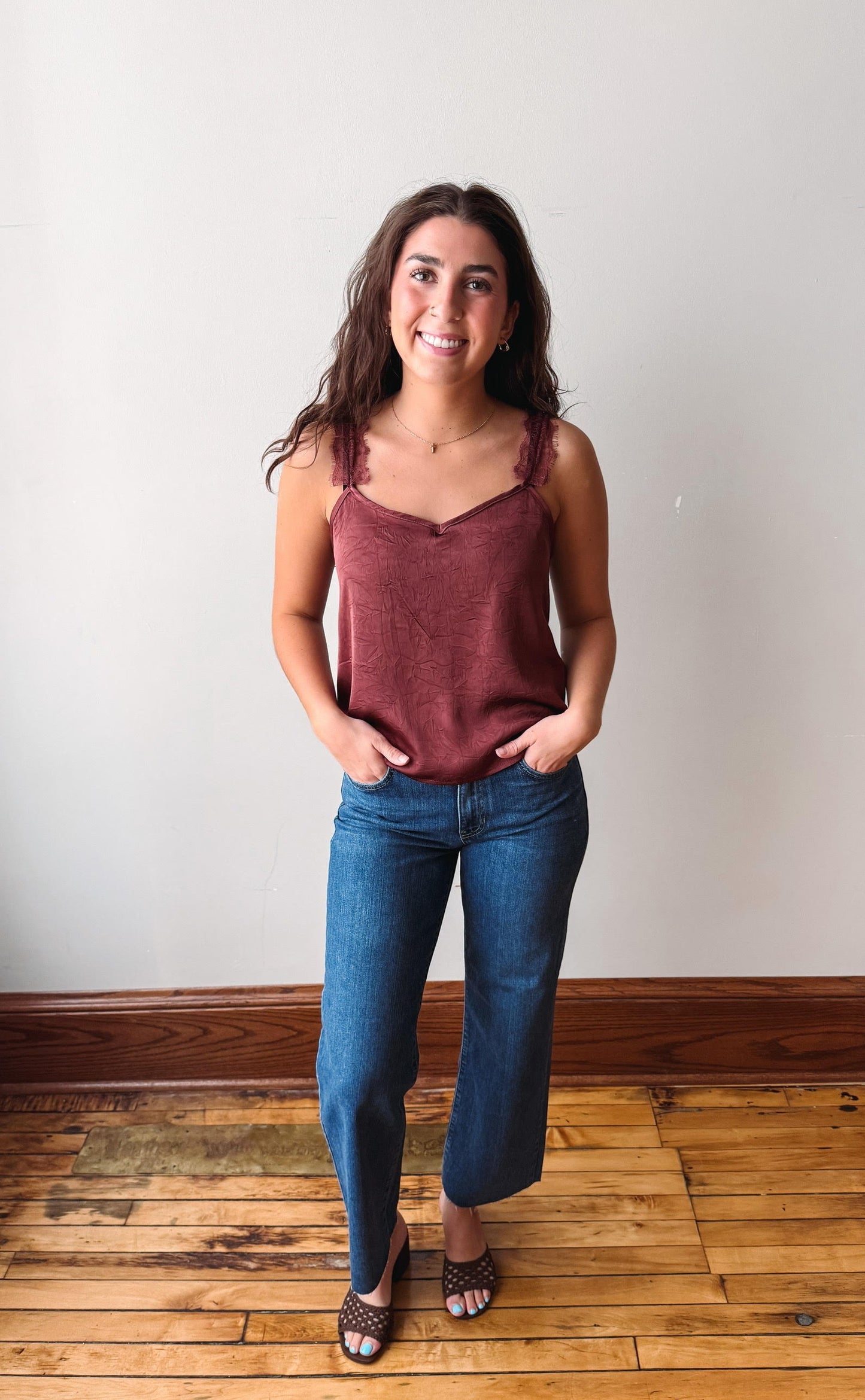 A woman stands indoors wearing a sleeveless dusty rose camisole top and blue jeans, with her hands in her pockets, against a plain off-white wall and wooden floor.