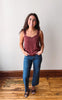 A woman stands indoors wearing a sleeveless dusty rose camisole top and blue jeans, with her hands in her pockets, against a plain off-white wall and wooden floor.