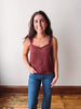 A woman modeling a rust-colored, textured camisole top with lace-trimmed spaghetti straps, paired with blue jeans, standing against a plain off-white wall with wooden flooring.