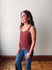 A woman wearing a rust-colored, textured camisole tank top and blue jeans, standing in a room with a light gray wall and wooden floor.